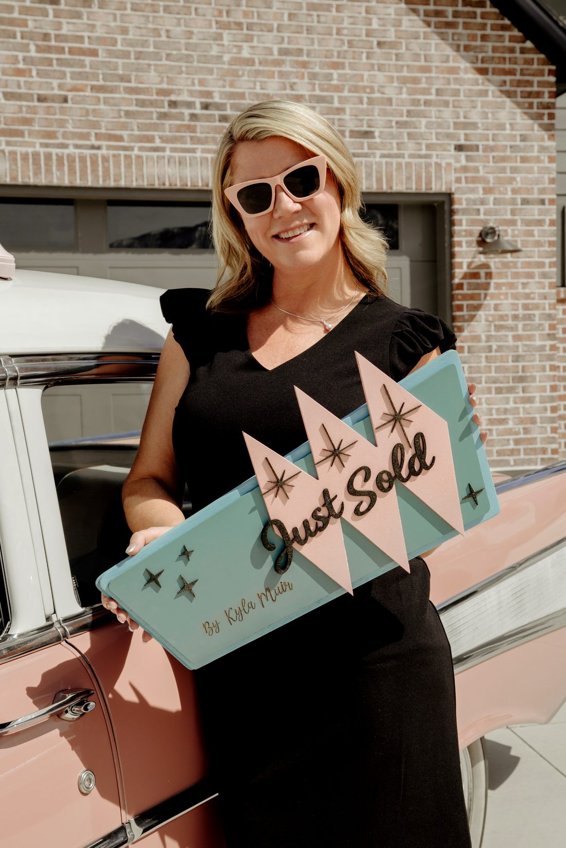 Kyla Muir holding a retro Just Sold sign beside her pink 1957 Bel Air